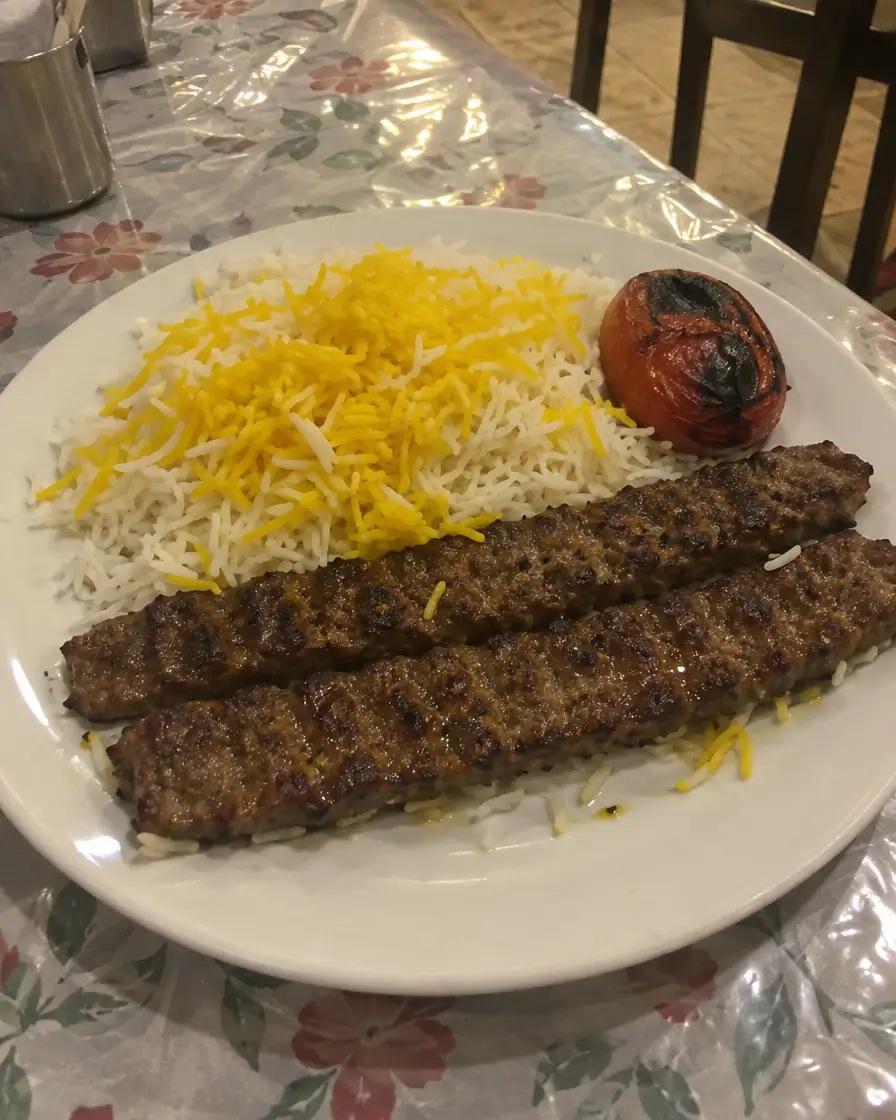 عکس گوشی از بشقاب چلوکباب کوبیده با برنج زعفرانی و گوجهی کبابی روی رومیزی پلاستیکی زیر نور زرد رستوران